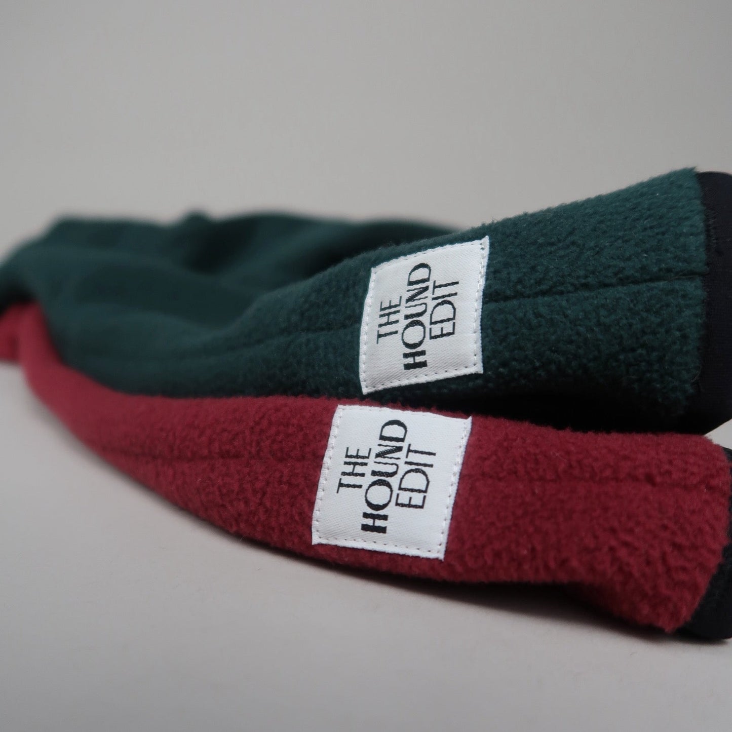 Close-up of two Italian Greyhound fleece jumpers in burgundy red and forest green with The Hound Edit brand label.