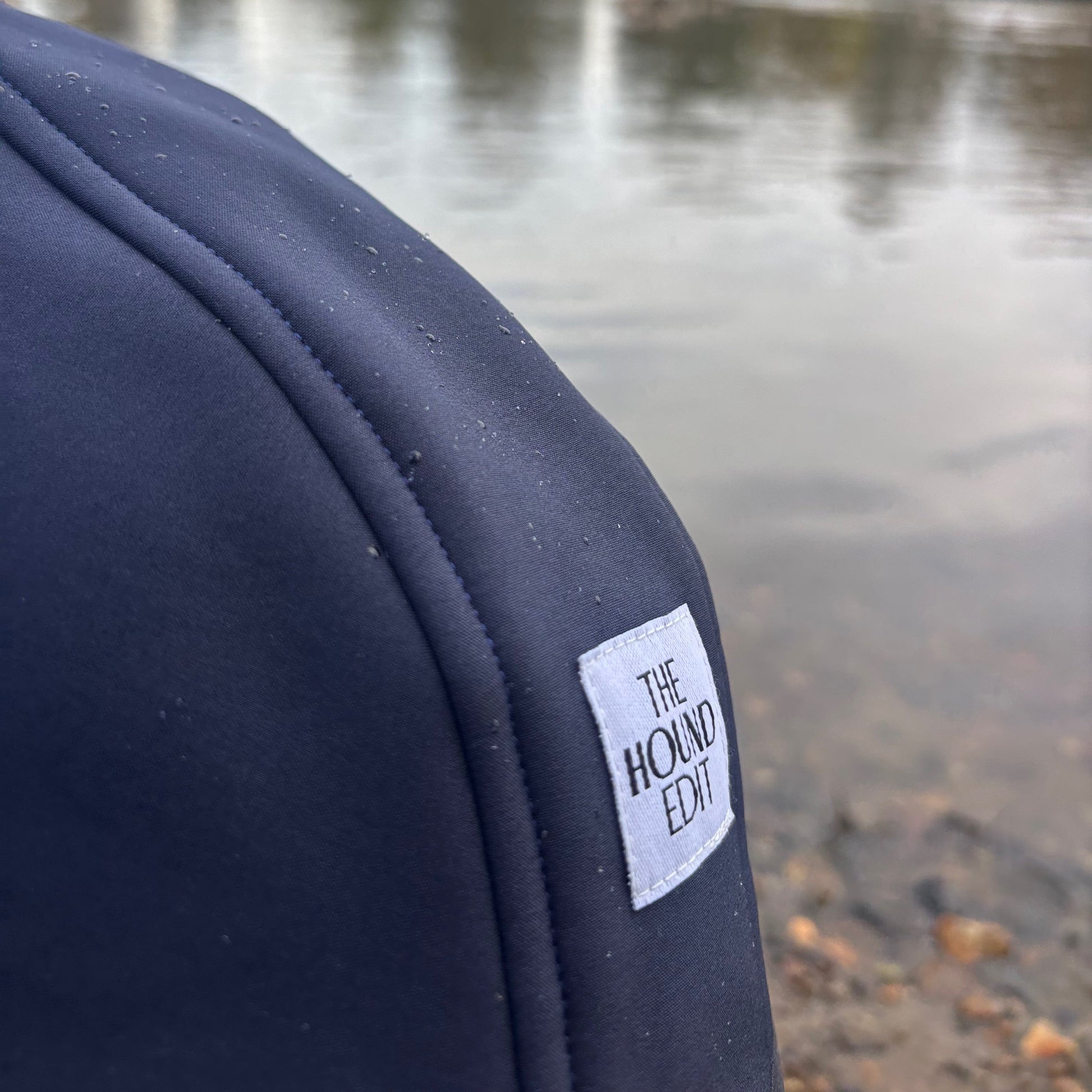Back of navy blue sleeveless Italian Greyhound coat outdoors in front of the river, showing stitching, raindrops, and The Hound Edit brand label
